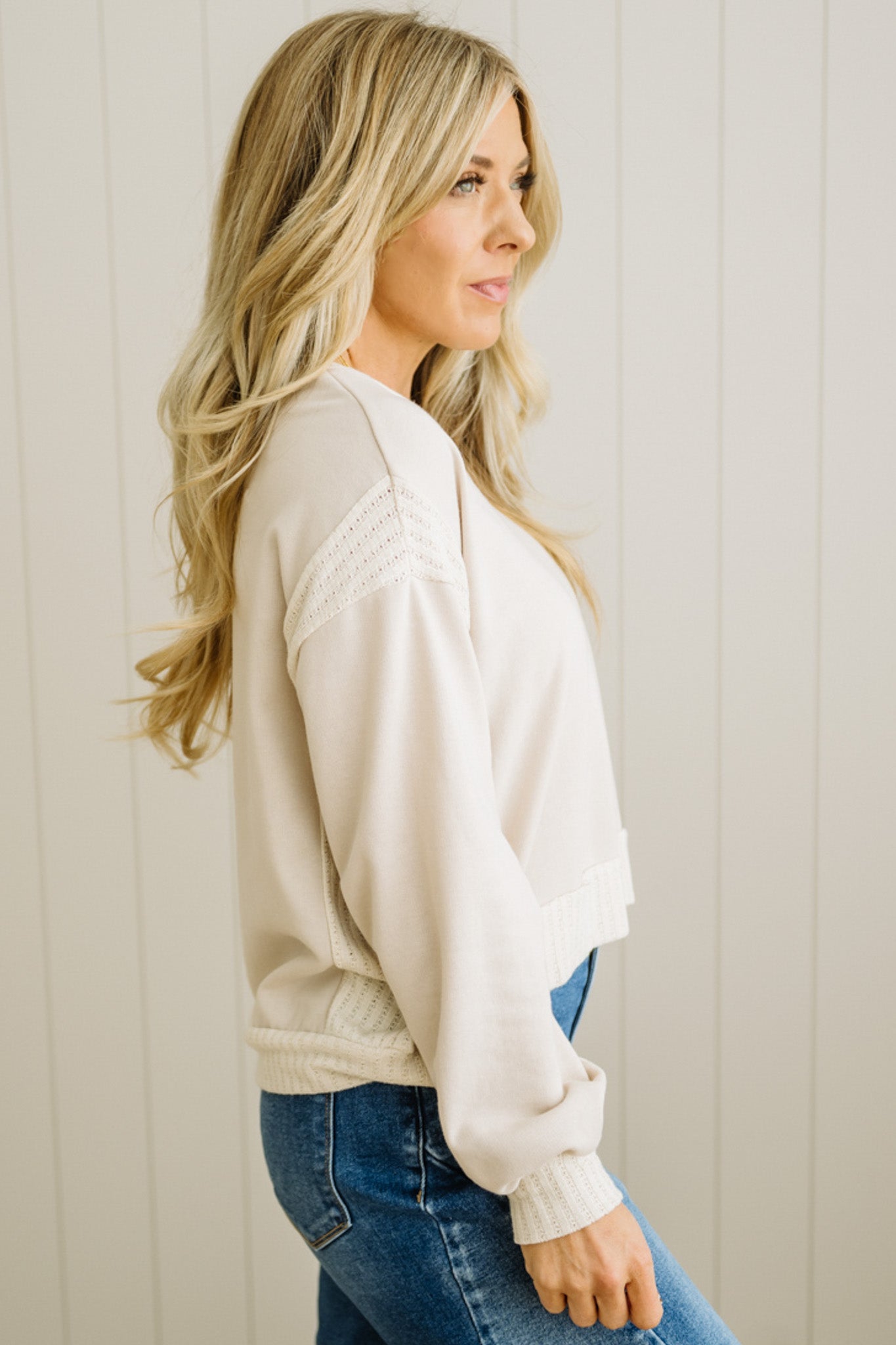 Woman wearing a beige sweater and blue jeans against a light wooden panel background