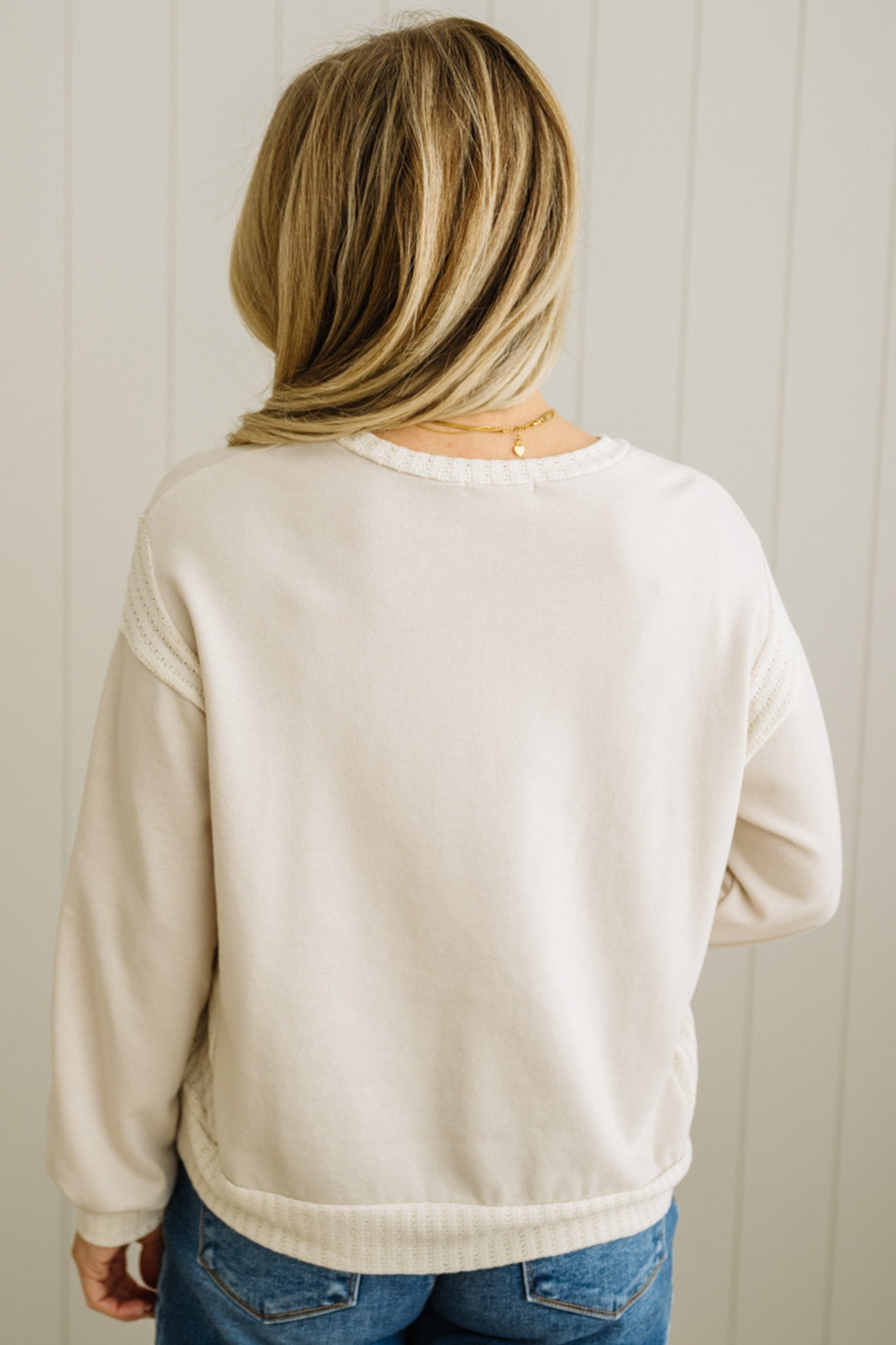 Person wearing a beige sweatshirt and blue jeans against a white paneled wall.