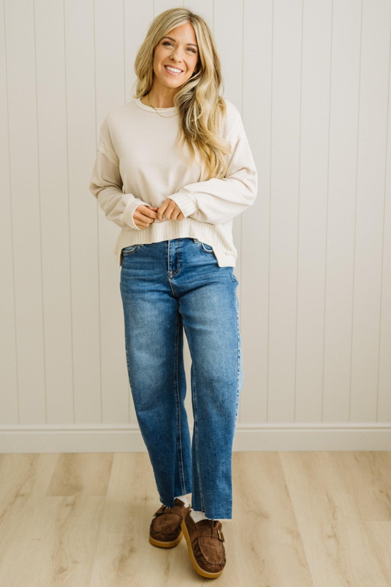 Woman wearing a white sweater, blue jeans, and brown shoes standing against a white paneled wall.
