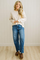 Woman wearing a white sweater, blue jeans, and brown shoes standing against a white paneled wall.