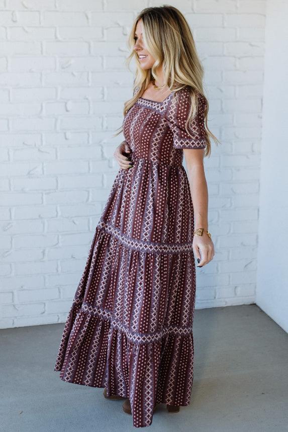 Woman wearing a long, patterned dress against a white brick wall.