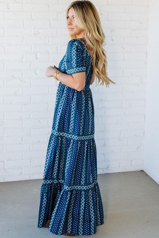 Woman wearing a blue patterned dress against a white brick wall.