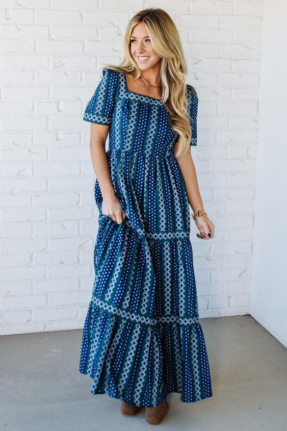 Woman wearing a blue patterned dress against a white brick wall.