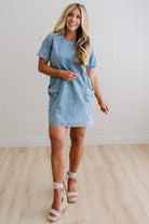 Blonde woman wearing a denim shift dress to the knee with short sleeves, side pickets and bow accents on each pocket.