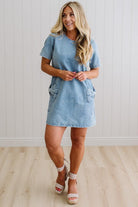 Blonde woman wearing a denim shift dress to the knee with short sleeves, side pickets and bow accents on each pocket.
