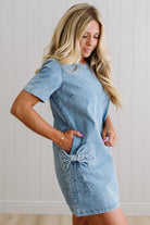 Blonde woman wearing a denim shift dress to the knee with short sleeves, side pickets and bow accents on each pocket.