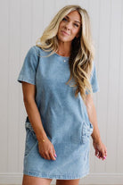 Blonde woman wearing a denim shift dress to the knee with short sleeves, side pickets and bow accents on each pocket.