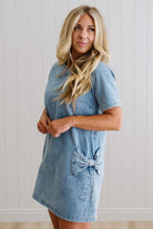 Blonde woman wearing a denim shift dress to the knee with short sleeves, side pickets and bow accents on each pocket.