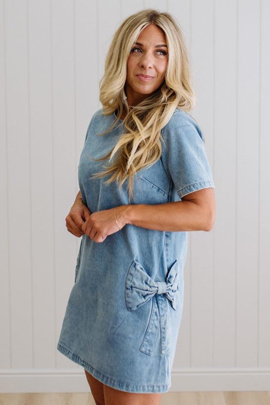 Blonde woman wearing a denim shift dress to the knee with short sleeves, side pickets and bow accents on each pocket.