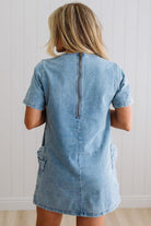 Blonde woman wearing a denim shift dress to the knee with short sleeves, side pickets and bow accents on each pocket.