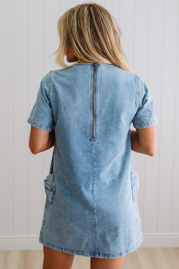 Blonde woman wearing a denim shift dress to the knee with short sleeves, side pickets and bow accents on each pocket.