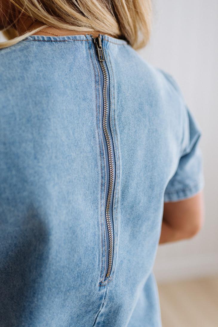Blonde woman wearing a denim shift dress to the knee with short sleeves, side pickets and bow accents on each pocket.