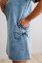 Blonde woman wearing a denim shift dress to the knee with short sleeves, side pickets and bow accents on each pocket.