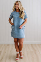 Blonde woman wearing a denim shift dress to the knee with short sleeves, side pickets and bow accents on each pocket.