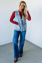 Woman wearing a denim vest over a red top with blue jeans against a white brick wall.