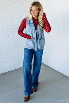 Woman wearing a denim vest over a red shirt with blue jeans against a white brick wall.