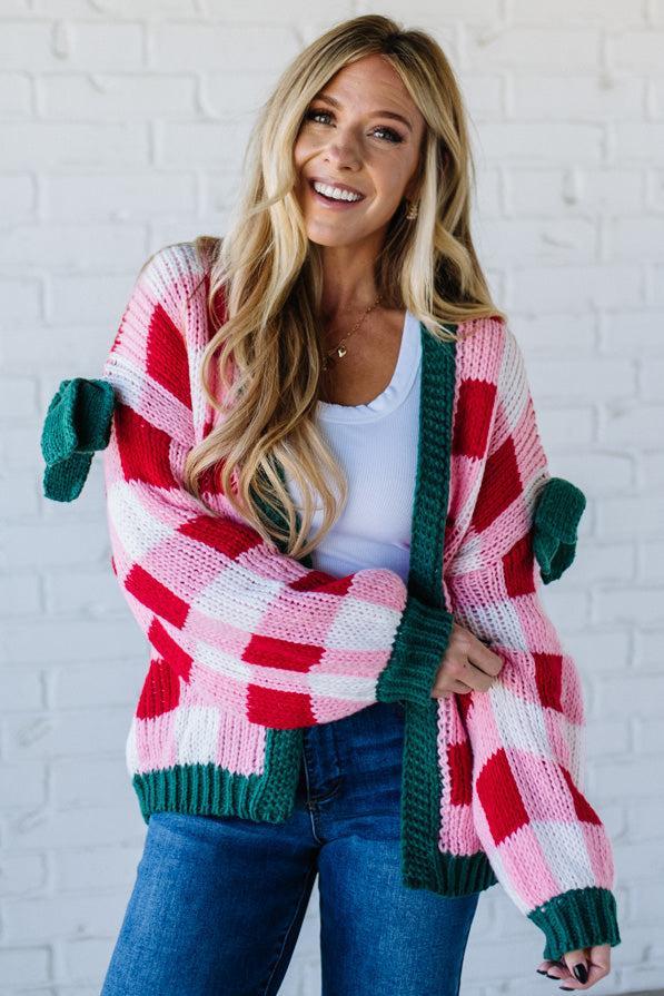 Woman wearing a colorful plaid cardigan with green accents against a white brick wall.