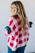 Person wearing a red and white checkered sweater with green accents against a white brick wall.