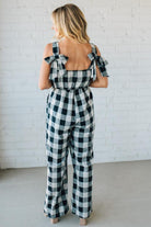 Woman wearing a black and white checkered jumpsuit with bow back straps against a white brick wall.