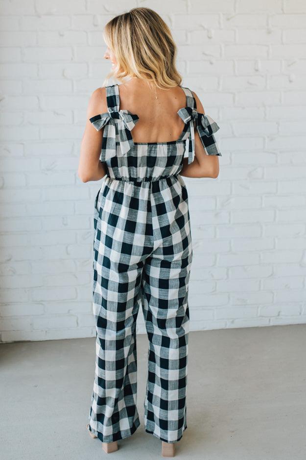 Woman wearing a black and white checkered jumpsuit with bow back straps against a white brick wall.