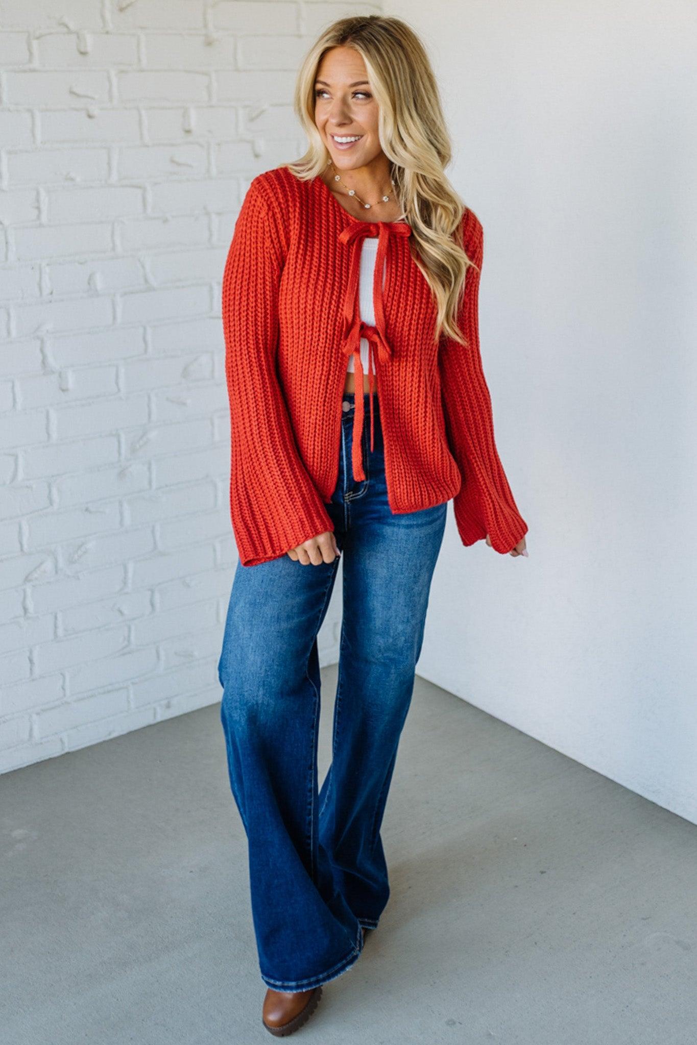 Woman wearing a red sweater and blue jeans standing against a white brick wall.