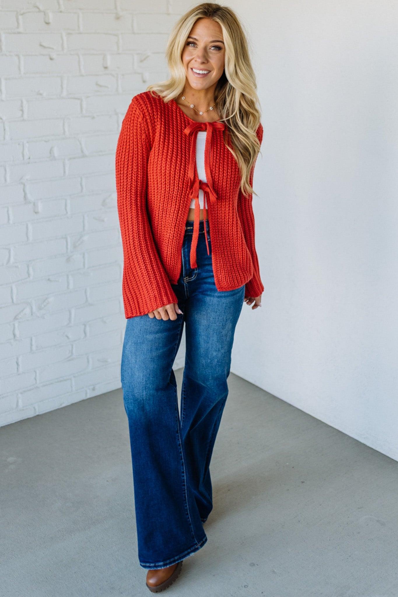 Woman wearing a red sweater and blue jeans standing against a white brick wall.