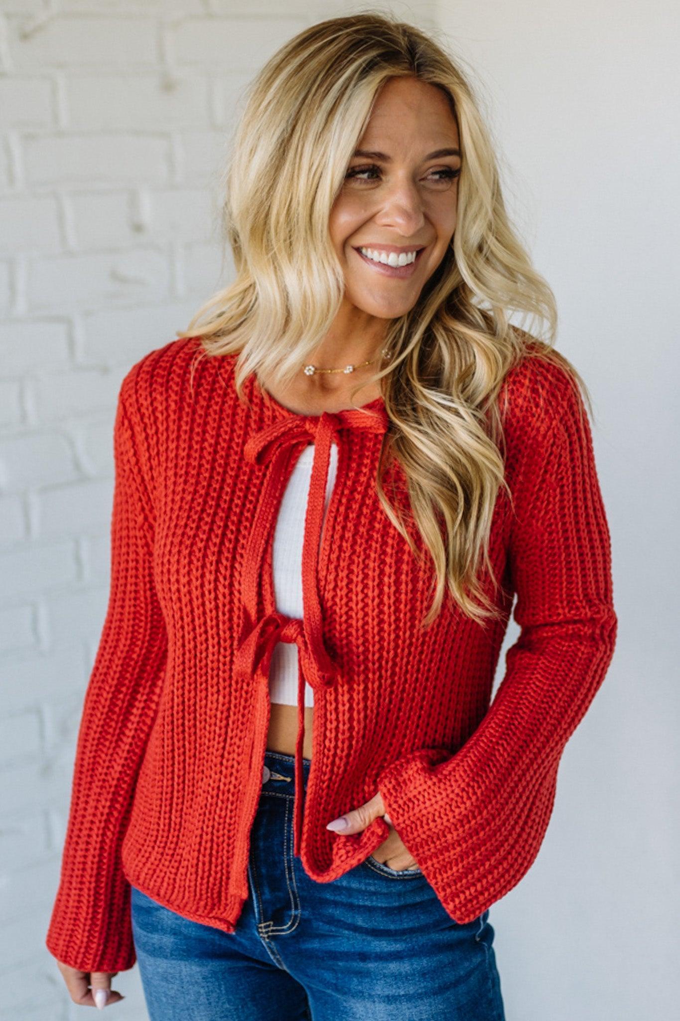 Woman wearing a red knitted cardigan over a white top and blue jeans against a white brick wall.