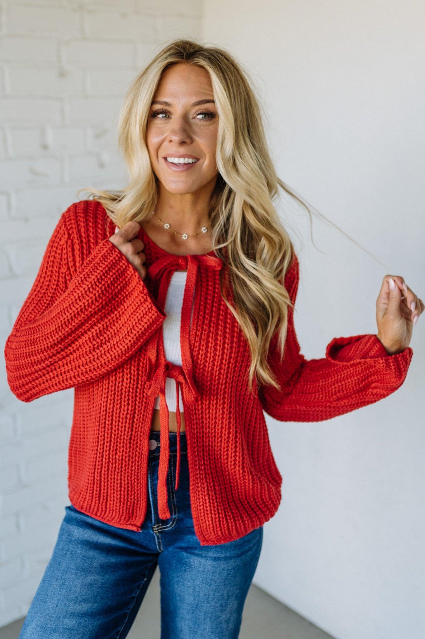 Woman wearing a red knitted cardigan over a white top with blue jeans against a white brick wall.
