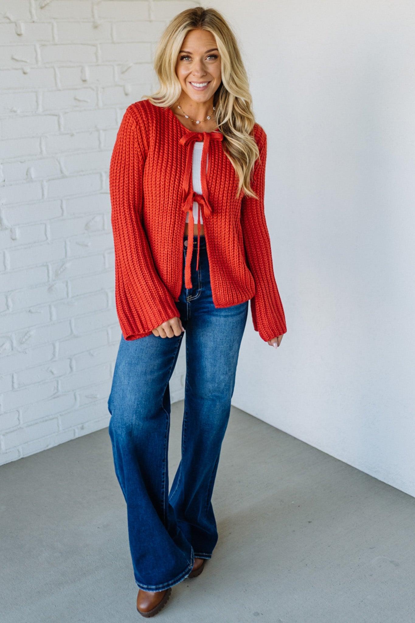 Woman wearing a red sweater and blue jeans against a white brick wall.