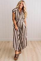 BLONDE WOMAN WEARING A SLEEVELESS WIDE LEG, WIDE STRIPE JUMPSUIT STANDING ON A LIGHT WOOD FLOOR AND NEUTRAL WALL BEHIND.