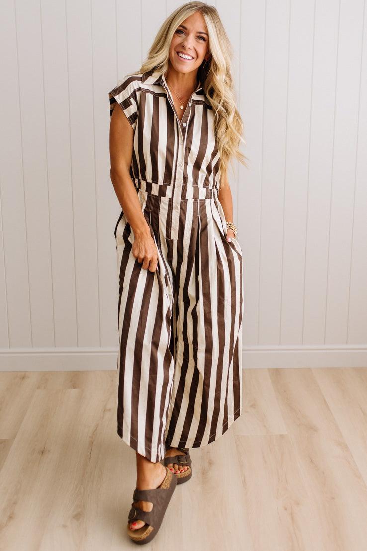 BLONDE WOMAN WEARING A SLEEVELESS WIDE LEG, WIDE STRIPE JUMPSUIT STANDING ON A LIGHT WOOD FLOOR AND NEUTRAL WALL BEHIND.