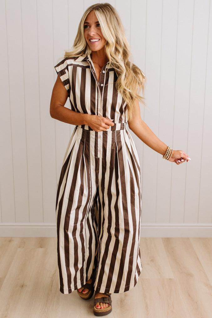 BLONDE WOMAN WEARING A SLEEVELESS WIDE LEG, WIDE STRIPE JUMPSUIT STANDING ON A LIGHT WOOD FLOOR AND NEUTRAL WALL BEHIND.