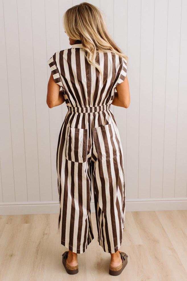 BLONDE WOMAN WEARING A SLEEVELESS WIDE LEG, WIDE STRIPE JUMPSUIT STANDING ON A LIGHT WOOD FLOOR AND NEUTRAL WALL BEHIND.