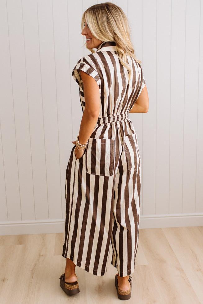 BLONDE WOMAN WEARING A SLEEVELESS WIDE LEG, WIDE STRIPE JUMPSUIT STANDING ON A LIGHT WOOD FLOOR AND NEUTRAL WALL BEHIND.