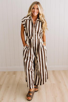 BLONDE WOMAN WEARING A SLEEVELESS WIDE LEG, WIDE STRIPE JUMPSUIT STANDING ON A LIGHT WOOD FLOOR AND NEUTRAL WALL BEHIND.