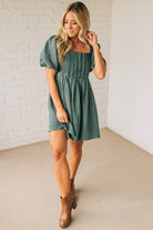Woman wearing a green dress with short sleeves against a white brick wall.