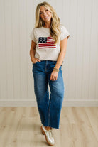 Woman wearing a t-shirt with an American flag design and blue jeans against a white wall.