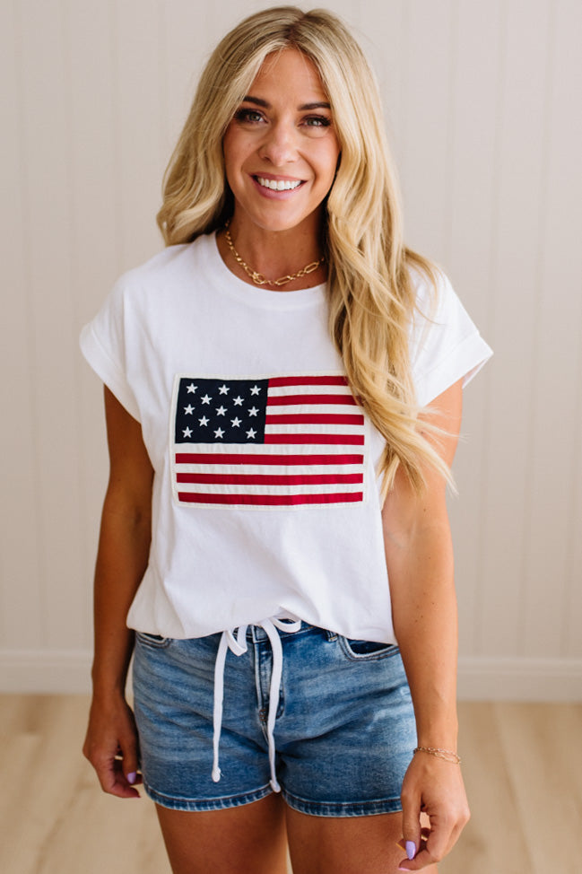 Woman wearing a white t-shirt with an American flag design and denim shorts indoors.