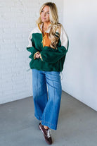 Woman wearing a green and white jacket with an orange shirt underneath, standing against a white brick wall.