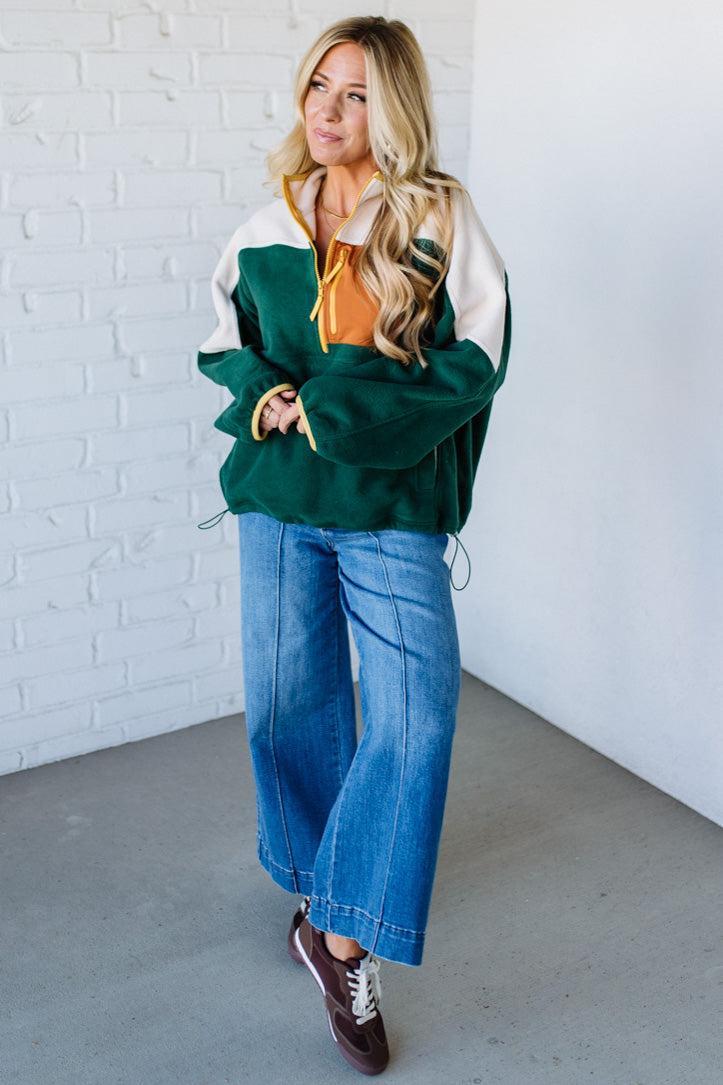 Woman wearing a green and white jacket with an orange shirt underneath, standing against a white brick wall.