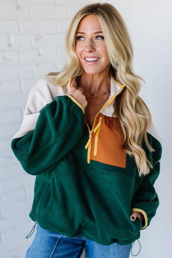Woman wearing a green and white jacket with a yellow zipper against a white brick wall.