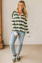 Woman wearing a green and white striped shirt with light blue jeans against a white brick wall.