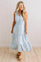 A woman modeling a sleeveless light blue tiered maxi dress with a smocked waist, paired with straw slide sandals, standing against a white paneled wall and light wood floor.