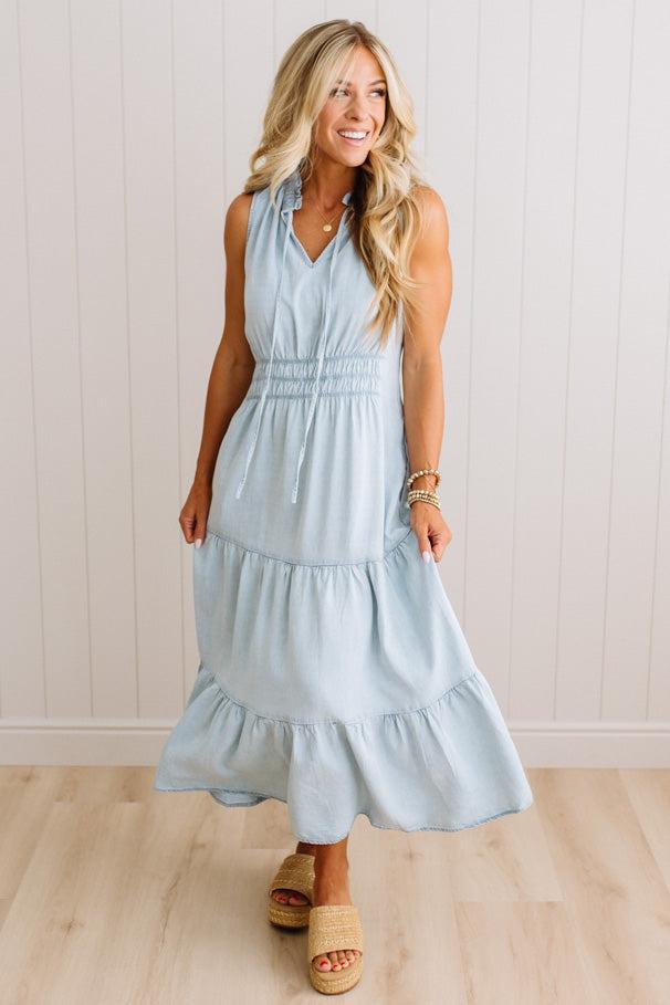 A woman modeling a sleeveless light blue tiered maxi dress with a smocked waist, paired with straw slide sandals, standing against a white paneled wall and light wood floor.