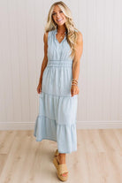 A woman modeling a sleeveless light blue tiered maxi dress with a smocked waist, standing on a light wood floor against a white paneled wall.