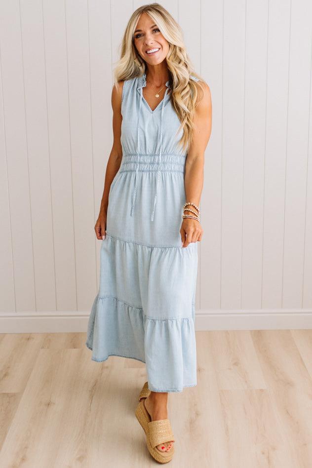 A woman modeling a sleeveless light blue tiered maxi dress with a smocked waist, standing on a light wood floor against a white paneled wall.
