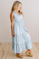 A woman wearing a light blue sleeveless tiered maxi dress with a smocked waist and side pockets, paired with woven tan wedge sandals, standing in front of a white paneled wall and light wood floor.