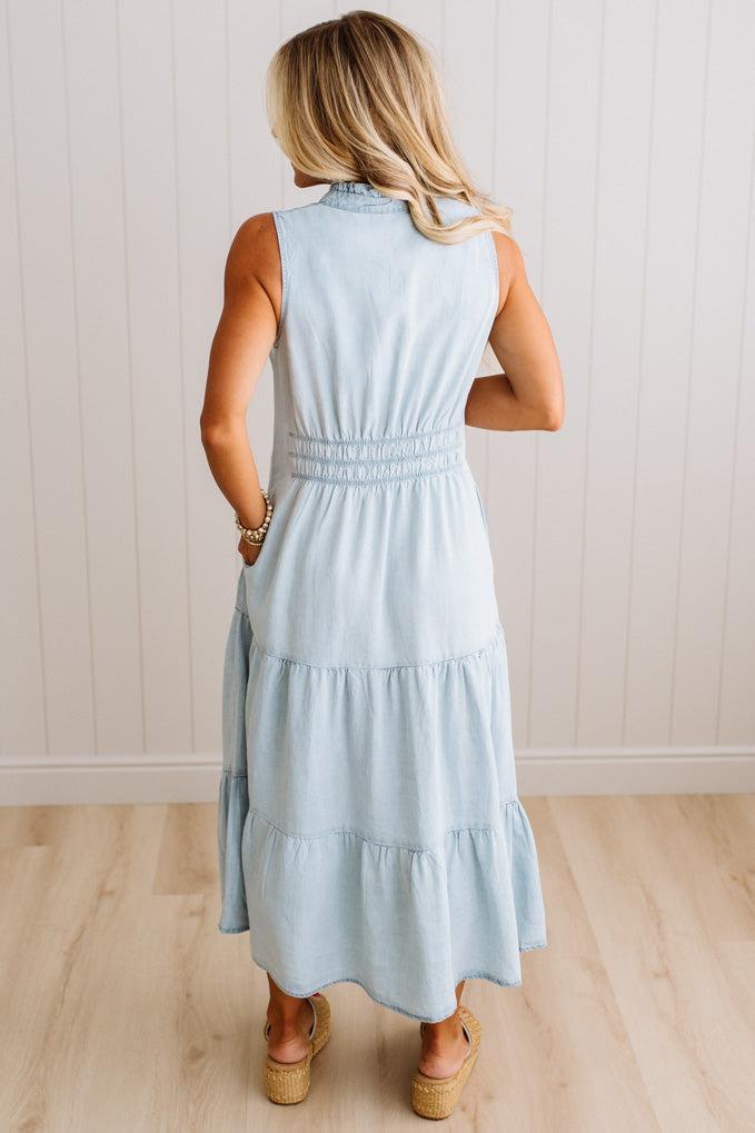 A woman viewed from behind wearing a sleeveless light blue tiered maxi dress with a smocked waist, standing on a light wood floor against a white paneled wall.