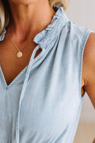 A close-up of a woman wearing a light blue sleeveless top with a ruffled collar and tie detail, paired with a delicate gold pendant necklace.