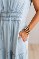 Close-up of a person wearing a light blue smocked dress, with one hand in a side pocket and stacked gold beaded bracelets on the wrist.